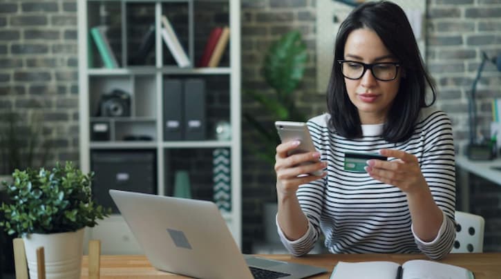 Mulher branca, de cabelos pretos, lisos e curtos usa óculos e blusa de meia manga listrada em tons branco e verde. Está sentada em frente a uma mesa com um notebook prata aberto, segurando um cartão de crédito na mão esquerda e um smartphone na mão direita.