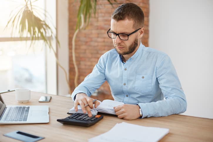 Homem sentado com um caderno, caneta, calculadora e notebook calculando as contas para o começo do ano.