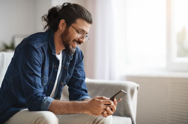 Homem de cabelo comprido, barba e óculos, vestindo uma jaqueta de jeans, sorrindo enquanto usa um celular em um ambiente bem iluminado.