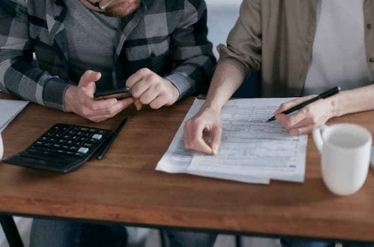 Duas pessoas sentadas à mesa analisam formulários e fazem cálculos, utilizando uma calculadora e celular, enquanto discutem como a alíquota do IPI sobre carros influencia no valor do imposto a ser pago.