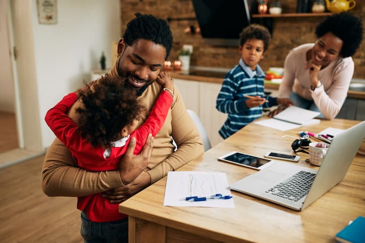 Pais e dois filhos estão sentados na mesa com papéis, canetas e notebook. Eles sorriem e se divertem enquanto estudam sobre como trabalhar educação financeira para crianças.