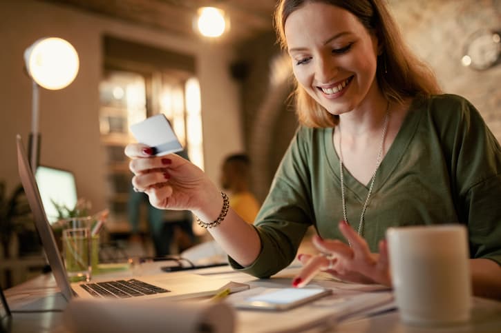 Mulher sentada à mesa com um notebook em sua frente e uma xícara de café ao lado. Ela segura o cartão de crédito em uma mão, enquanto paga a conta no seu celular. Ela está feliz por entender o que é inadimplente no Serasa e quitou suas dívidas.