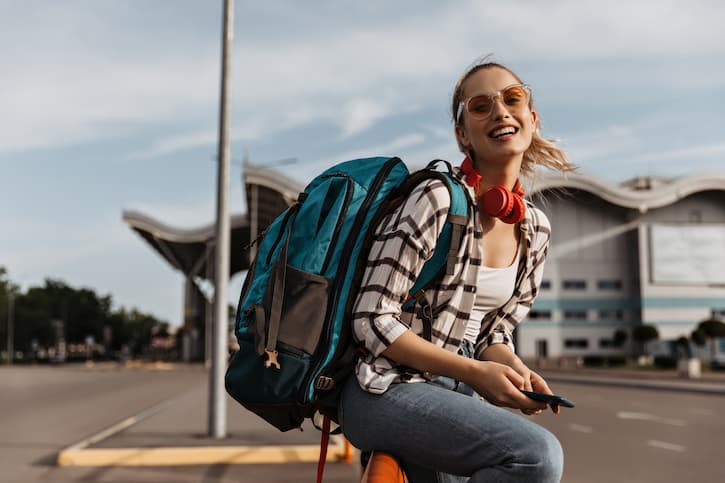 Mulher em frente ao aeroporto preparada para viajar com uma mochila nas costas e sentada em sua mala. Ela sorri para a foto.