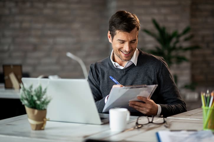 Homem sentado à mesa em frente ao computador com uma prancheta e caneta na mão. Ele está lendo sobre Fundo de Investimento, o que é e suas vantagens e agora está conhecendo as opções.