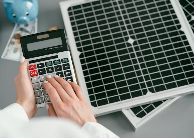 Mulher segura calculadora em frente a uma mesa com um cofre de porquinho, cédulas de dinheiro e moedas e duas placas fotovoltaicas. Ela entendeu sobre energia fotovoltaica o que é, seu preço e busca como pagar.