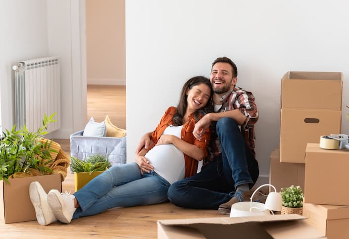 Casal sentado no chão da sala de um apartamento garden, cercado por caixas e objetos da mudança, com plantas ao redor, aproveitando o momento em seu novo lar via consórcio de apartamentos.