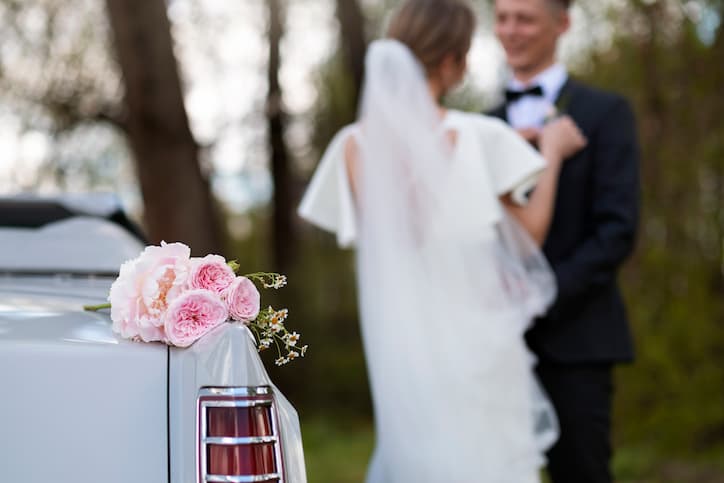 Buquê de flores cor-de-rosa apoiado em carro clássico durante as bodas de casamento, com noiva de véu e noivo em traje elegante desfocados ao fundo