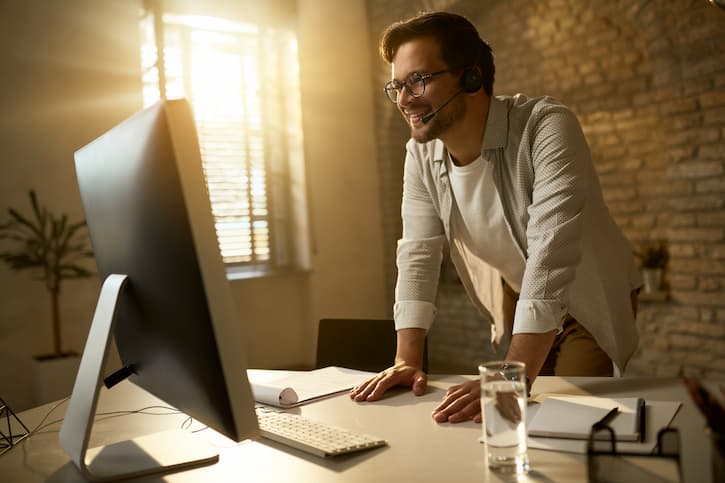 Homem sorridente em frente ao computador do seu escritório em casa. Ele usa um fone de ouvido e na mesa estão papéis e um copo d’água. 