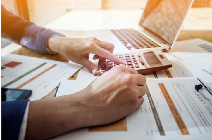 Mãos humanas estão debruçadas sobre papéis que parecem contas, a mão direita segura uma caneta e anota em um dos papéis, enquanto a mão esquerda digita em uma calculadora sobre a mesa.