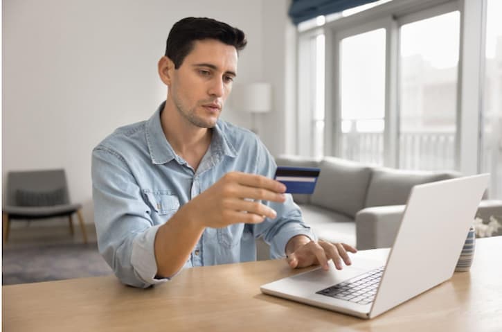 Homem de cabelo escuro, com expressão séria, segurando um cartão de crédito na mão e olhando para a tela do laptop em uma sala bem iluminada.